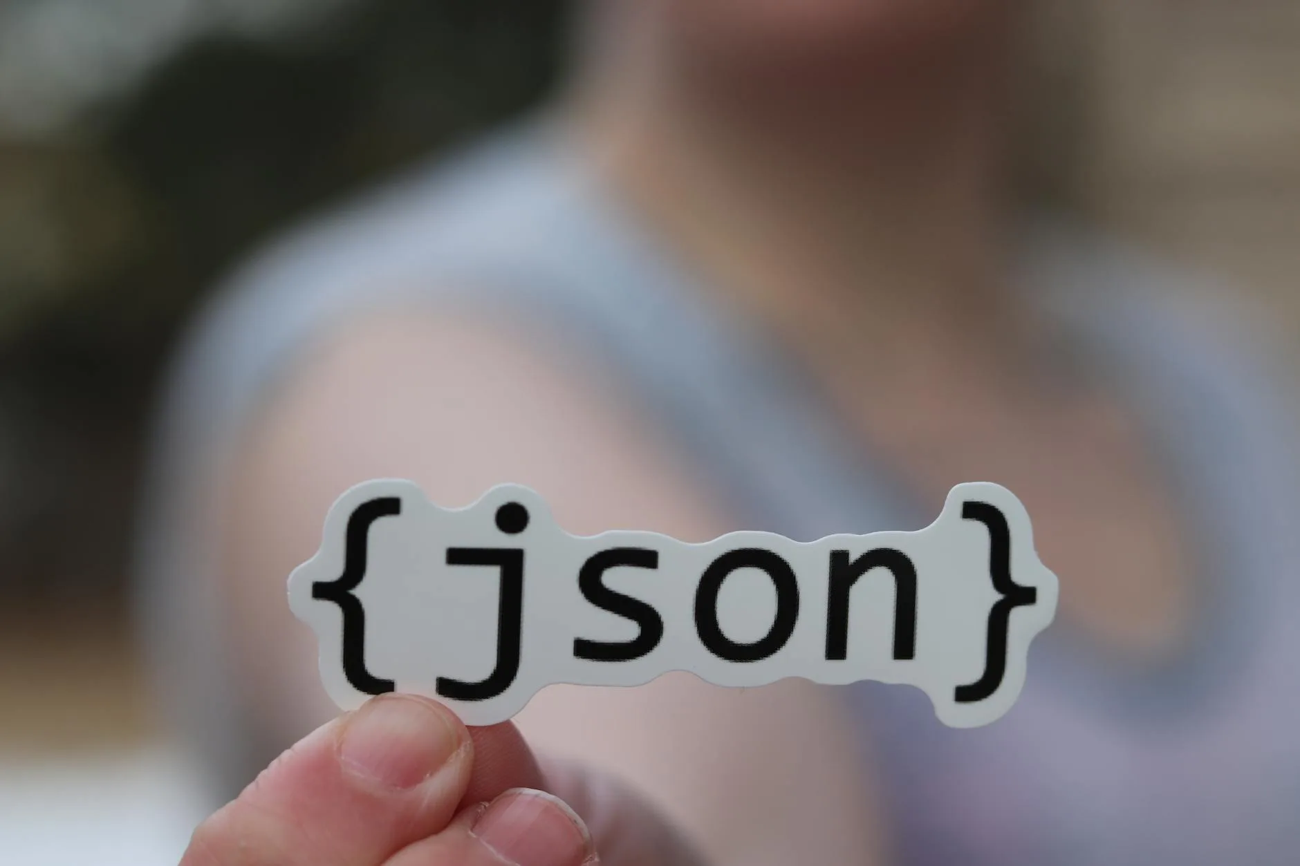 A hand holding a die-cut sticker printed with the JSON logo in curly braces, held up in front of a blurred background.